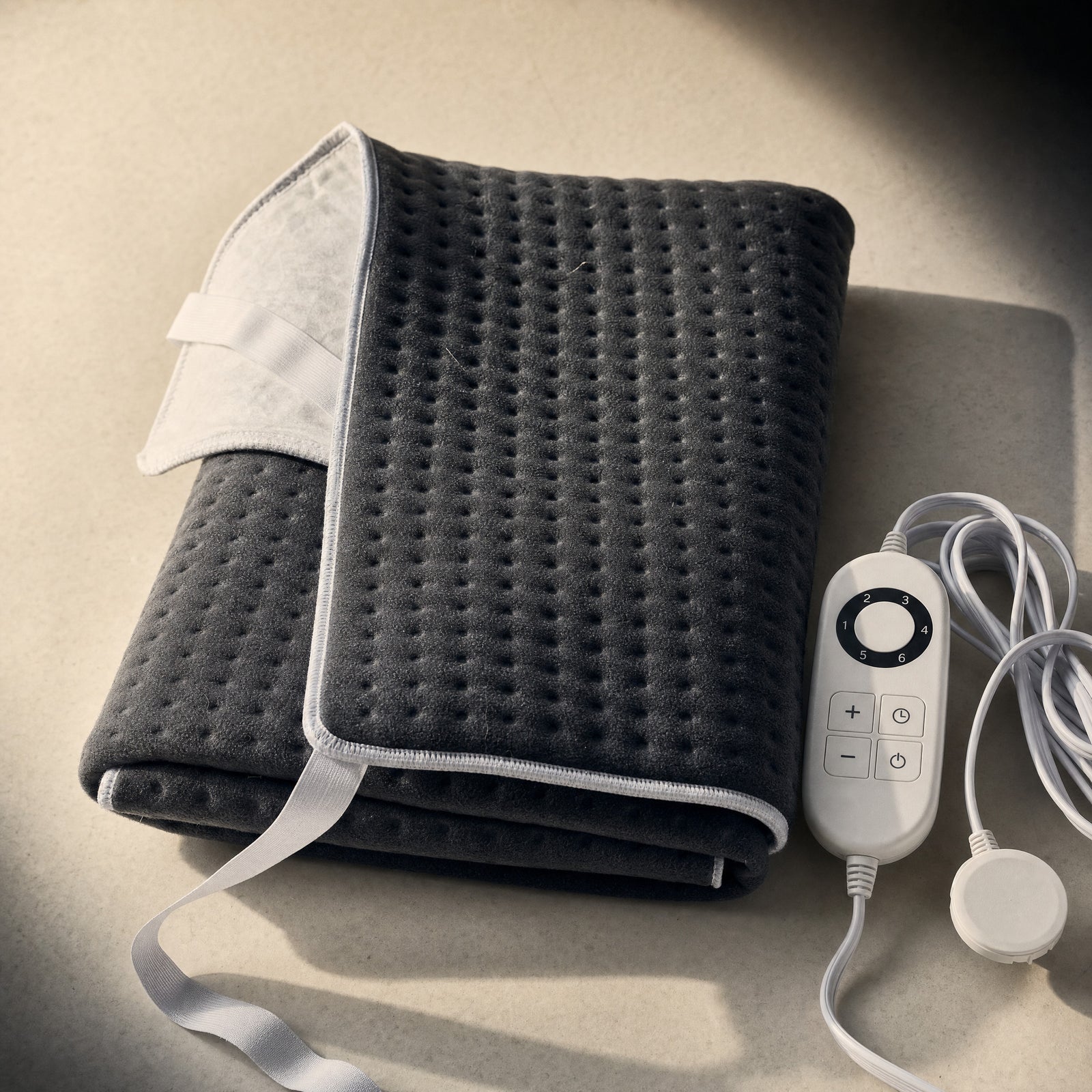 Charcoal Rowan single heated underblanket folded on a warm off-white surface, stamp-welded dimple quilting on the fleece face, one corner turned back to show the pale underside and its white six-setting controller beside it