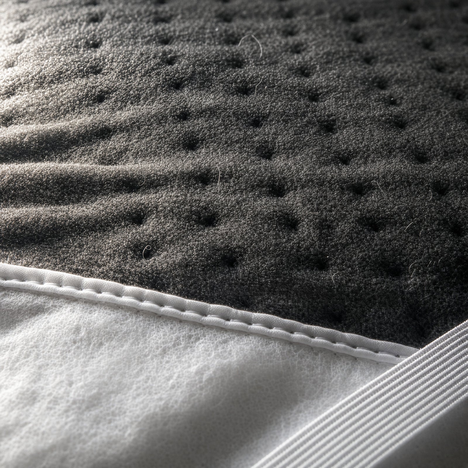 Close-up of the edge of the Rowan double heated underblanket, charcoal polyester fleece top face with its stamp-welded dimples above the bound seam, the plain white non-woven underside and one white elastic corner strap below