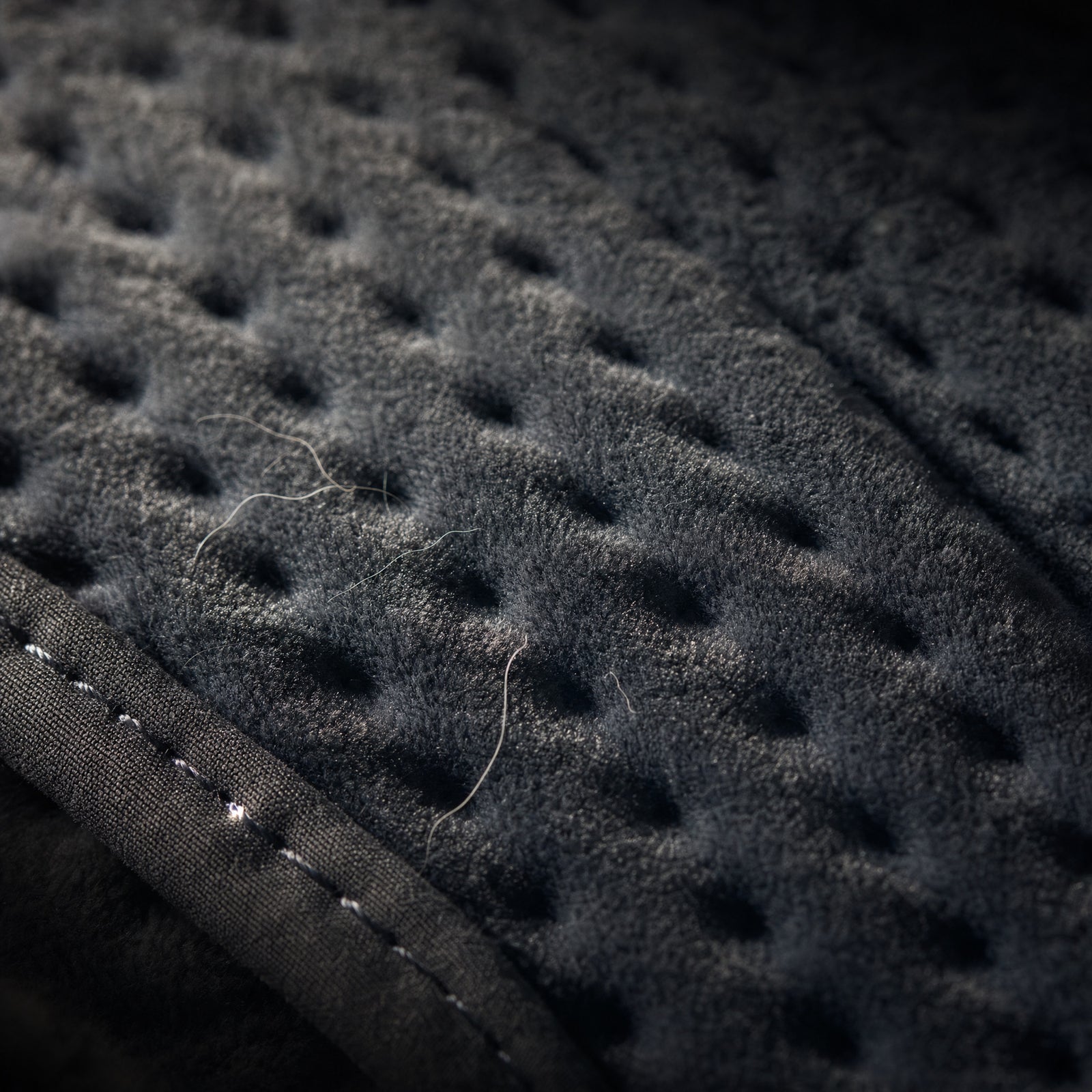 Close-up of the quilted face of the Rowan heated cushion, charcoal polyester fleece with even rows of stamp-welded dimples and the plain bound edge crossing the corner, raking light picking out loose fibres
