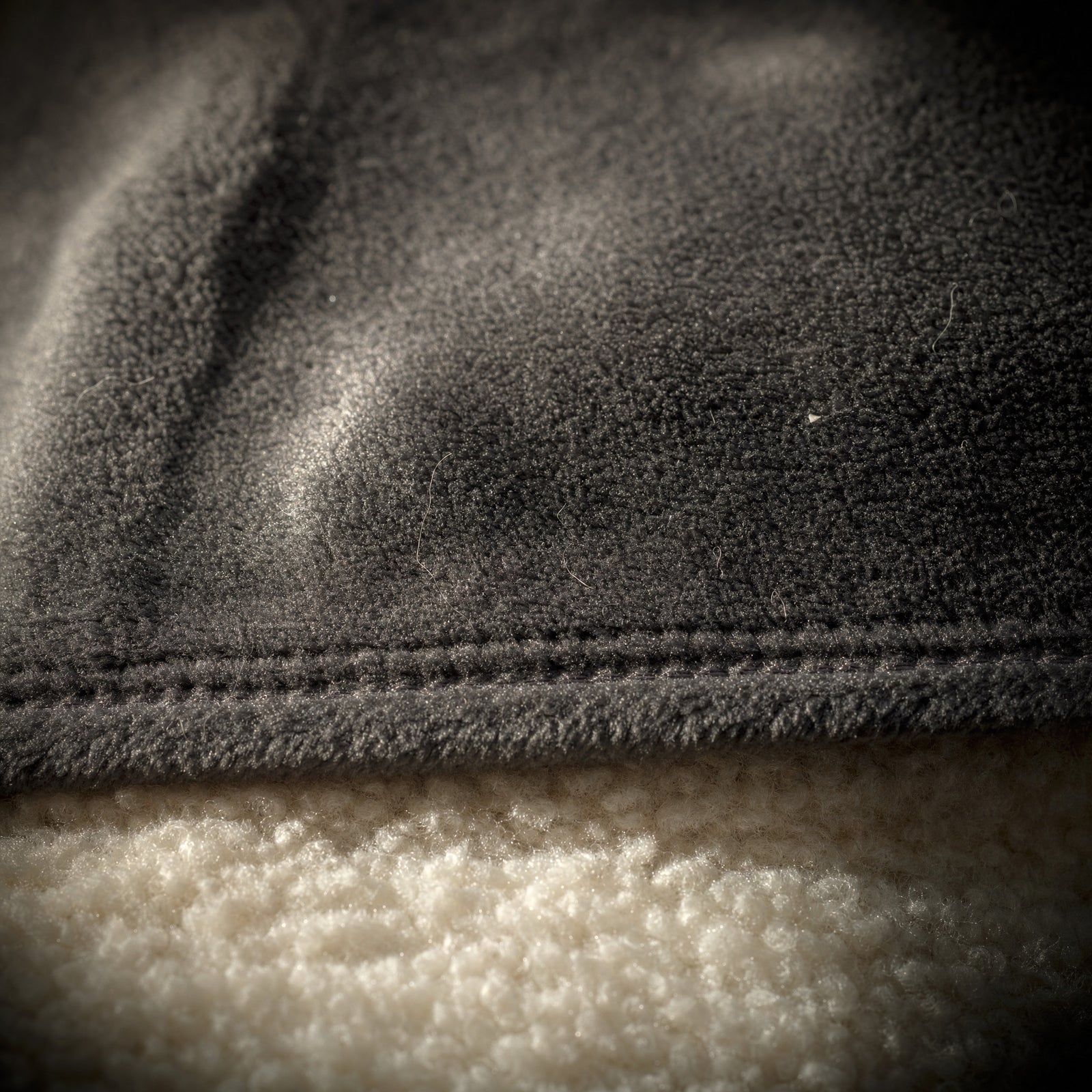 Close-up of the stitched hem of the Hazel heated throw, brushed charcoal polyester flannel face above and cream polyester sherpa reverse below, raking light showing the short flat pile direction and a few loose fibres