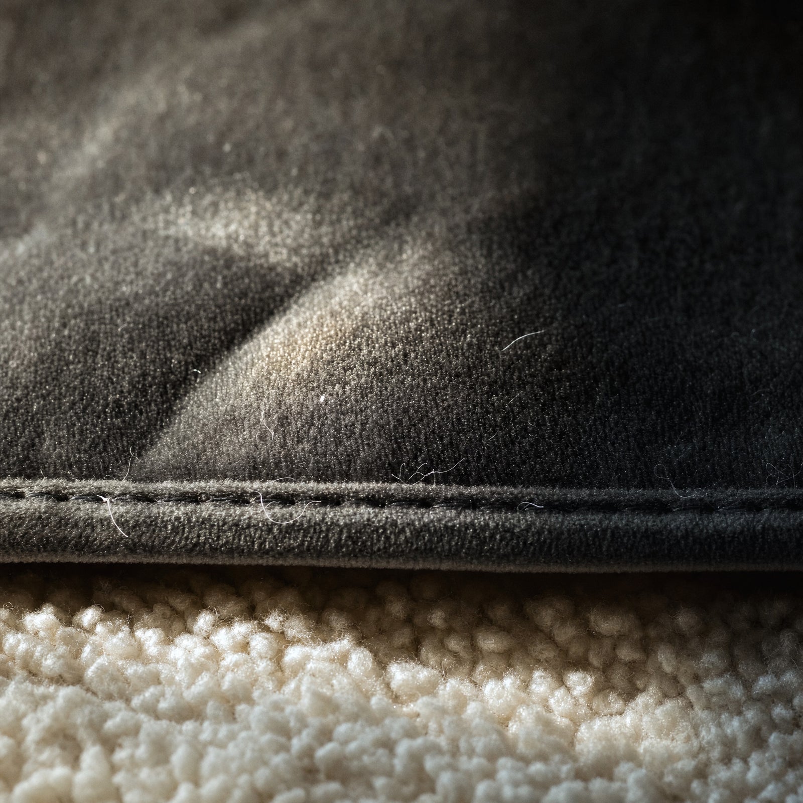 Close-up of the stitched hem of the Oak heated throw, brushed charcoal polyester flannel face above and cream polyester sherpa reverse below, raking light showing the short flat pile direction and a few loose fibres